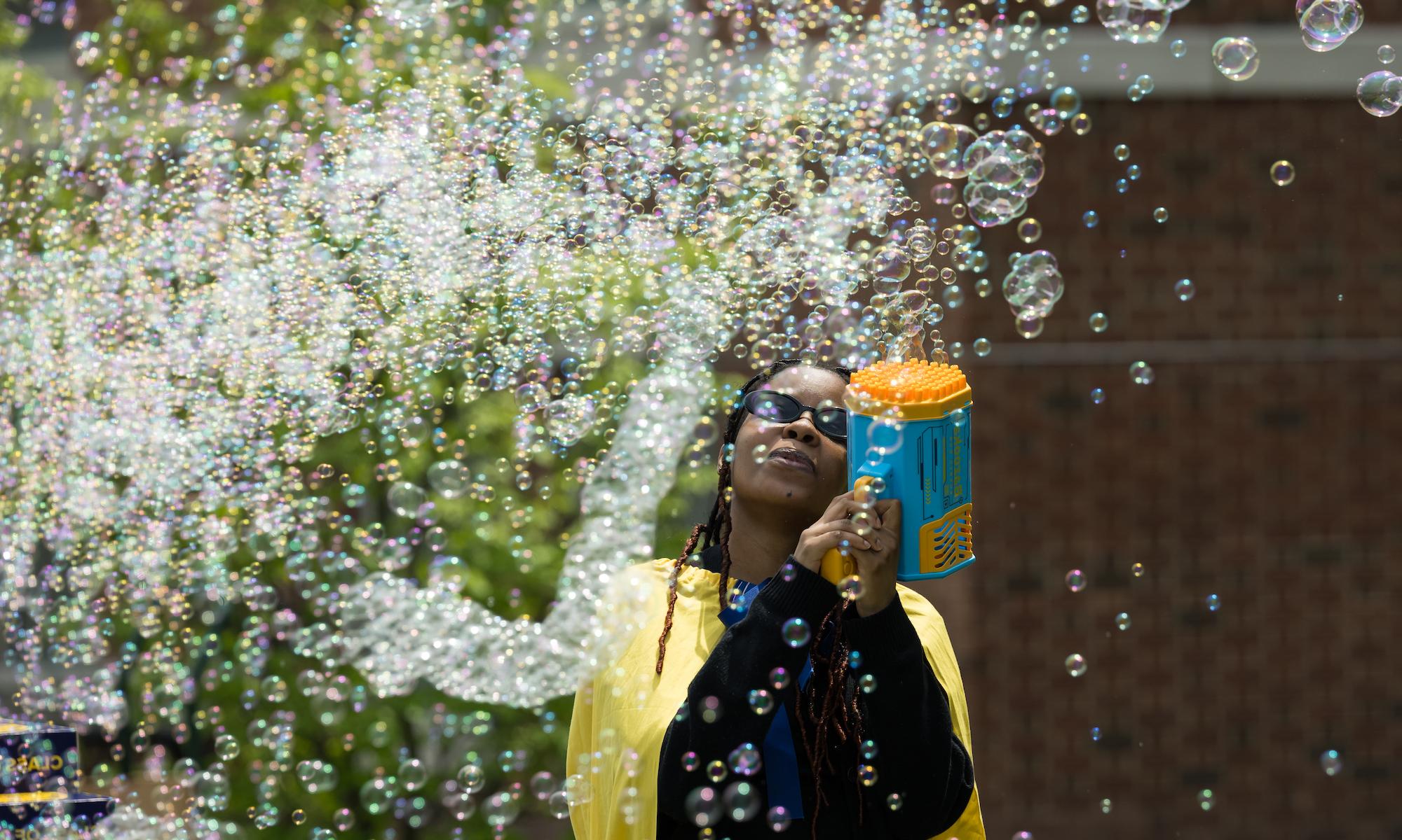 Student in graduation regalia surrounded by bubbles coming out of a bubble gun