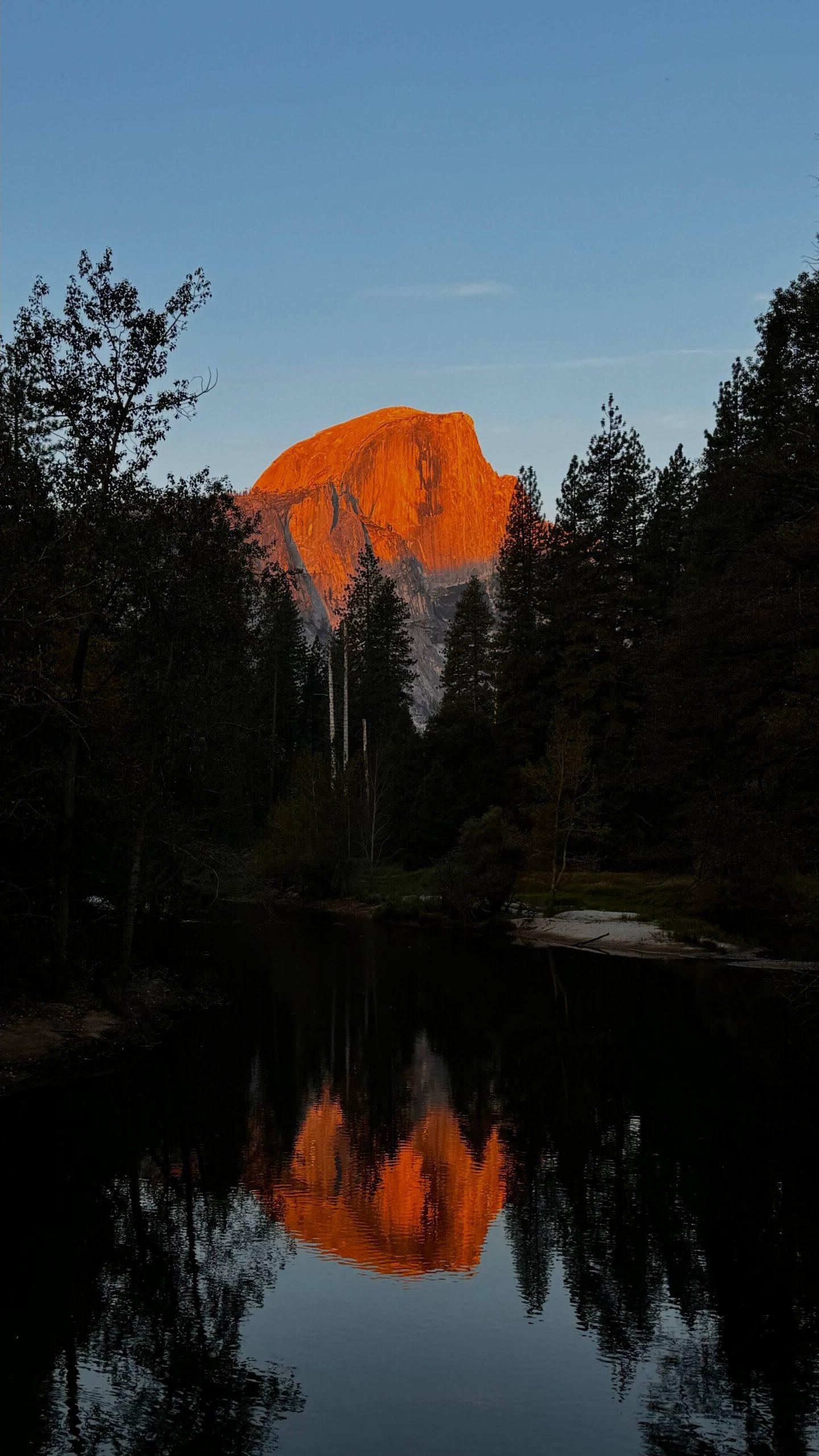 耀眼的橙色日落照亮了约塞米蒂国家公园的半圆顶山峰, framed by tall trees. The clear, still water below mirrors the vibrant rock formation against a deepening blue sky.