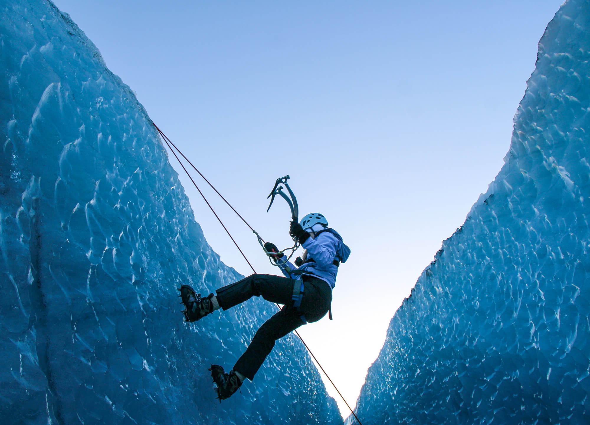 A person wearing climbing gear, including a helmet and crampons, is ice climbing between two tall ice walls. 他们使用冰镐并用绳索固定. The sky in the background is clear and bright.