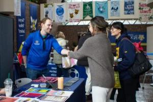 Individual wearing a blue U of R quarter zip hoodie tabling at a Wellness Fair smiling and greeting two students and shaking one of their hands. (Flourish Festival 2024)